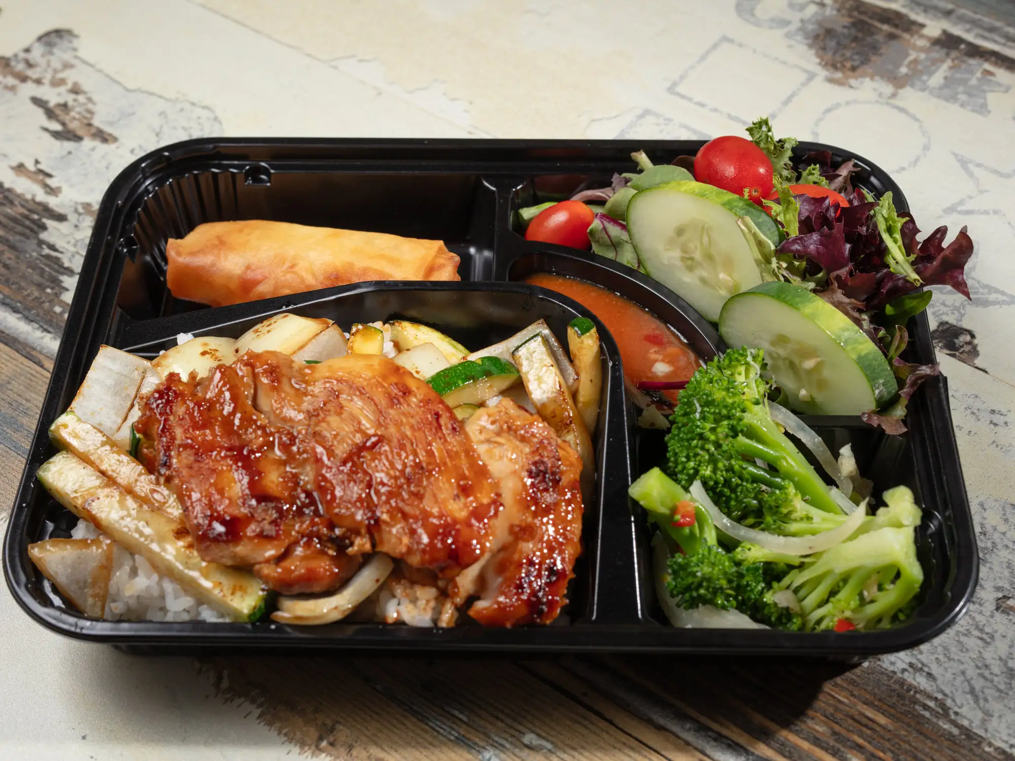 A black bento tray holding glazed chicken over grilled onion and zucchini, a spring roll, and a salad of mixed leaves, cucumber, tomato and broccoli