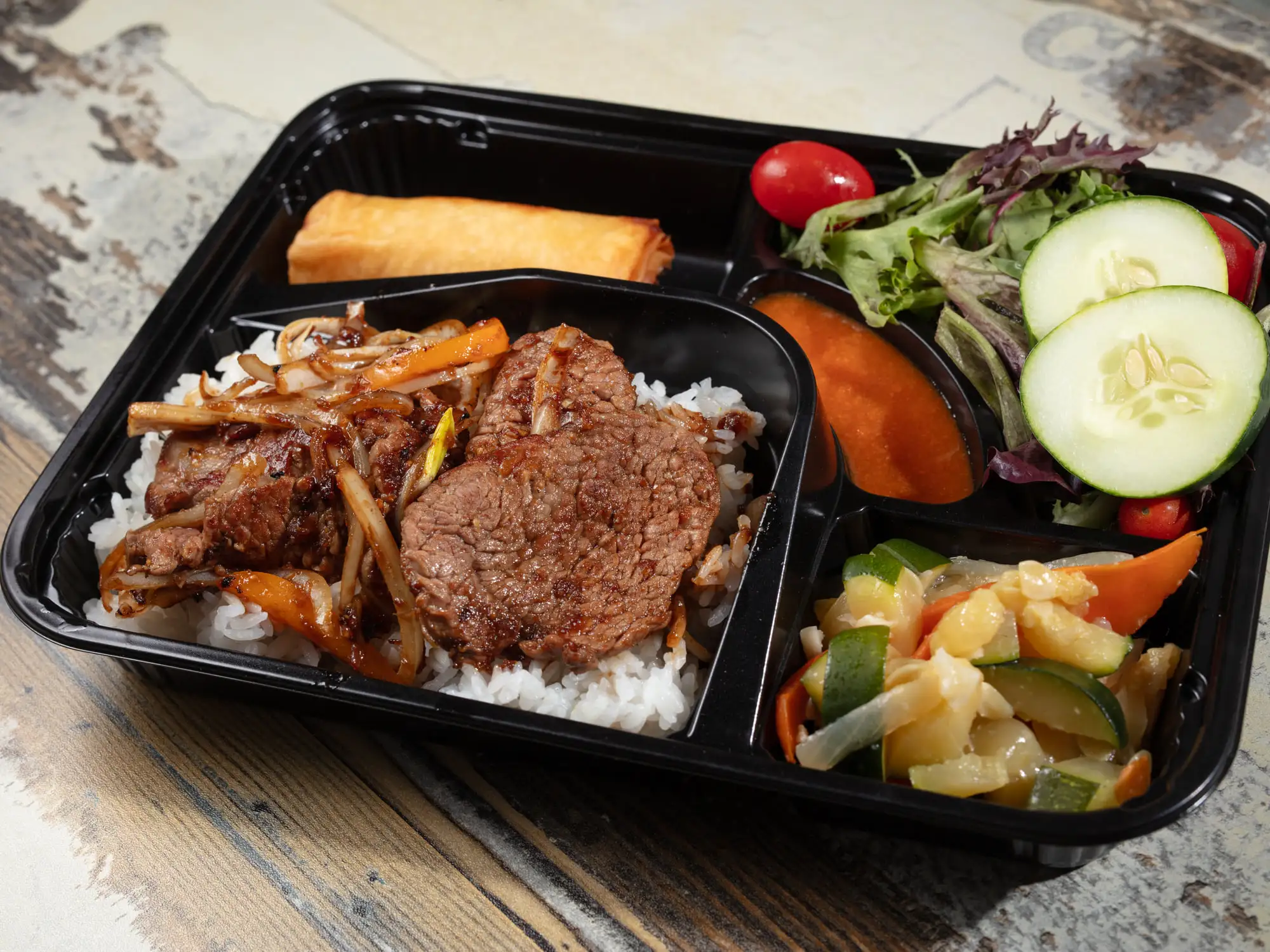 A black bento tray holding sliced beef with onion and carrot over white rice, a spring roll, grilled vegetables, and a salad of mixed leaves, cucumber, tomato and carrot